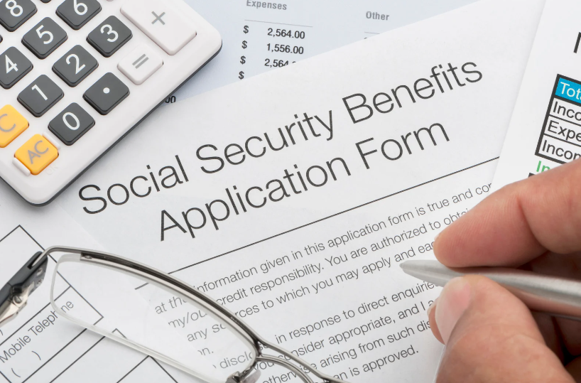 A person filling out a Social Security Benefits application form with a pen, surrounded by a calculator, eyeglasses, and financial documents.